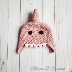 a pink crocheted hat with black eyes and teeth on top of a white wooden surface