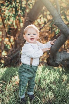 a little boy standing in the grass holding onto a tree branch