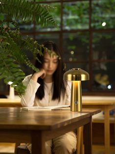 a woman sitting at a table with a lamp on top of it next to a plant