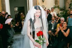 a woman in a white wedding dress and veil holding a red rose while people take pictures