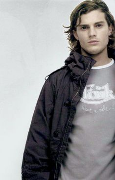 a young man with long hair wearing a black jacket and gray t - shirt standing in front of a white background