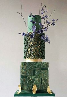 a three tiered green and gold wedding cake with flowers on top, sitting on a table