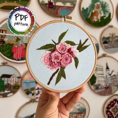 a hand holding up a embroidery hoop with pink flowers on it and many other embroidered pictures in the background