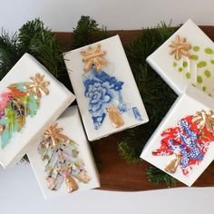 four ceramic boxes with designs on them sitting on a wooden tray next to pine branches