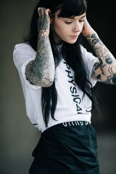 a woman with long black hair and tattoos on her arms, holding her hand to her head