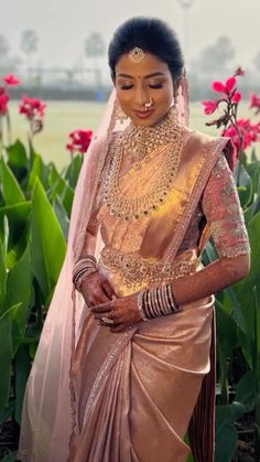 a woman in a pink and gold sari with jewelry on her head standing next to flowers