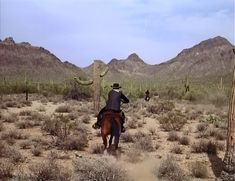 a man riding on the back of a brown horse through a desert landscape with mountains in the background