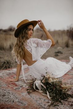 a woman in a white dress and brown hat sitting on a rug with her hands behind her head