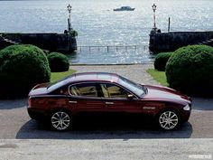 a maroon car is parked in front of some bushes near the water and a boat