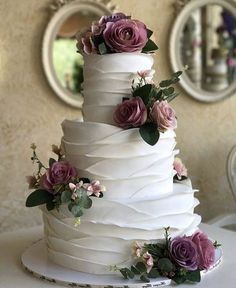 a three tiered white cake with purple flowers on the top and bottom, sitting on a table in front of a mirror