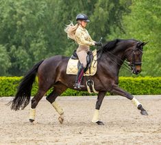 a woman riding on the back of a brown horse across a dirt field with trees in the background