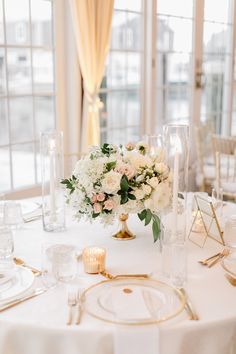 a white table topped with lots of flowers and gold place settings next to tall windows