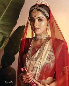 a woman in a red and gold bridal outfit with jewelry on her head, holding a green plant