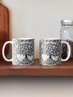 two coffee mugs sitting next to each other on a wooden shelf near a jar