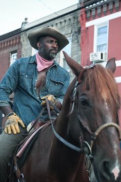 two men riding horses in front of a red building and one man wearing a cowboy hat