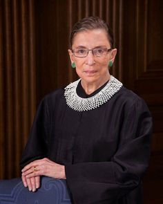 an older woman wearing glasses standing in front of a wooden paneled wall and holding a blue chair