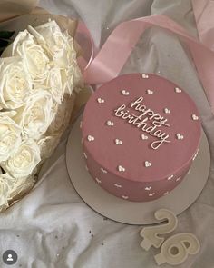 a pink birthday cake next to a bouquet of white roses and a heart shaped box