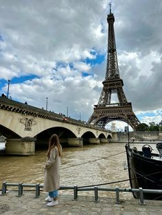 a woman is standing in front of the eiffel tower and boat on the river