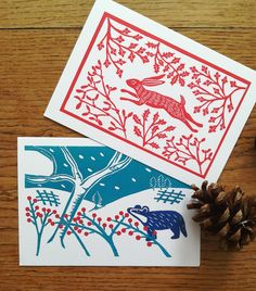 two cards with red and blue designs on them next to a pine cone, scissors and thread