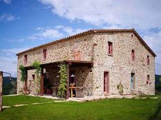 an old stone building with red shutters on the front and side windows, surrounded by lush green grass