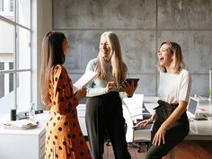 three women are laughing and having conversation in an office setting with large windows on the wall