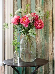 pink flowers in a glass vase sitting on a table next to a wooden wall with peeling paint