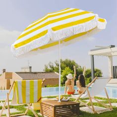 two women sitting in lawn chairs under an umbrella next to a swimming pool and deck chair