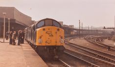 a yellow train traveling down tracks next to a loading platform with people standing on it