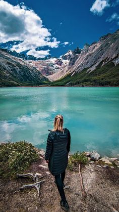 a woman standing on the edge of a lake looking at mountains and clouds in the sky