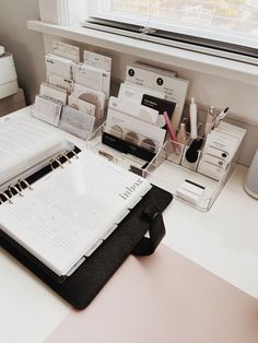 an office desk with several binders, pens and papers on it in front of a window