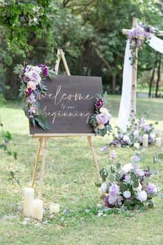 a welcome sign with purple flowers and greenery on the grass at a wedding ceremony