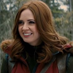 a woman with long red hair smiles at the camera while standing in front of trees