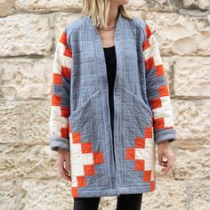 a woman standing in front of a stone wall wearing an orange and white jacket with squares on it