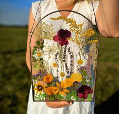 a woman holding up a purse with flowers on the front and side, while standing in a field