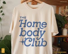 "The Homebody Club" Unisex T-shirt Design by jojoyu This item is made to order. If you want an oversized fit please size up! for reference, the model on the photo is usually a medium but wearing XL for an oversized fit. About the shirt: ◦ color: ivory ◦ relaxed fit  ◦ soft-washed, garment-dyed fabric ◦ double-needle stitching throughout the tee makes it highly durable  ◦ made with medium fabric (6.1 oz/yd²) consisting of high quality, 100% ring-spun US cotton for long-lasting comfort ◦ made with Shirt With Quotes, Fall Tees For Women, Women's Tshirt, Trendy Vinyl Shirt Ideas, Shirt Styles, Popular Tshirt Designs 2024, Cute Shirt Ideas Vinyl, Tshirt Inspiration, Shirt Graphics