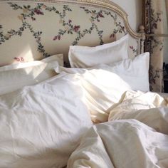 an unmade bed with white linens and floral wallpaper on the headboard