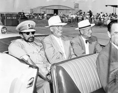 four men in suits and hats are sitting on a car with other people around them