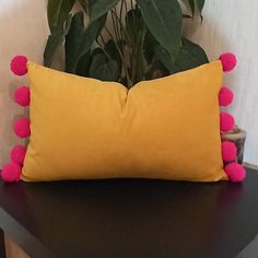 a yellow pillow with pink pom - poms sits on a black table next to a potted plant