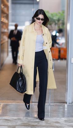 a woman in a yellow coat and black pants is walking down the street with a handbag