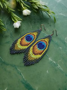 a pair of beaded peacock earrings sitting on top of a green table next to flowers