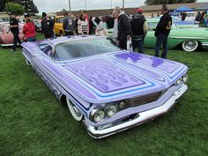 an old purple car parked on top of a lush green field next to other cars