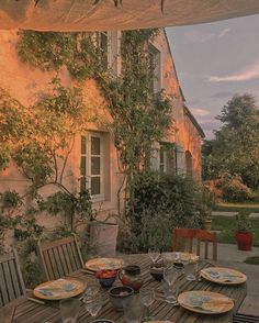 an outdoor dining table with plates and glasses on it, in front of a house