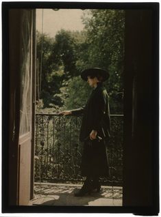 a woman in a long coat and hat standing on a balcony looking out the window