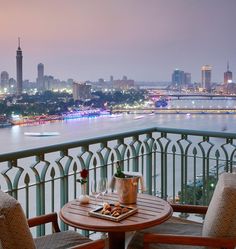 two chairs and a table on a balcony overlooking the water with city lights in the background