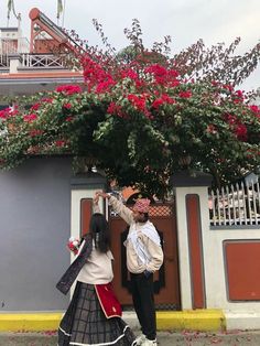 two people standing in front of a house with flowers on the roof and one person reaching up