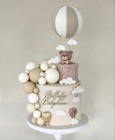 a white cake with balloons and a teddy bear sitting on top of it in front of a gray background