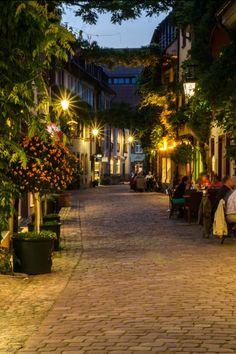 a cobblestone street with people sitting at tables and lights on the buildings in the distance