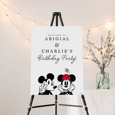 a mickey and minnie mouse birthday party sign on an easel with lights in the background