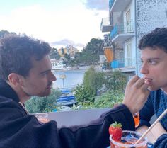 two men sitting at an outdoor table eating food and drinking drinks with chopsticks in their mouths