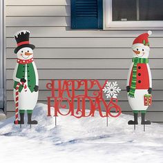 two snowmen standing next to each other in front of a house with the word happy holidays written on it